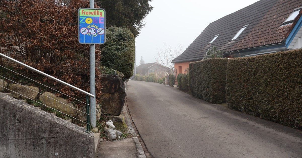 An empty street with a speed limit sign indicating 20, 30, and 40 km/h. A house is on the right with a gray roof and windows.