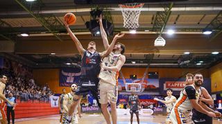 Basketball players compete in an indoor arena. One player jumps to dunk while another tries to block. The audience watches from the stands.