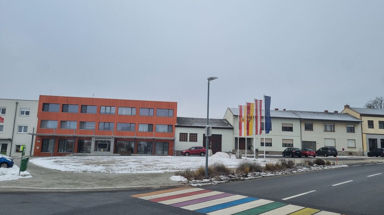 Eine Außenszene mit einem roten Gebäude und einem weißen Gebäude mit Flaggen davor. Schnee liegt auf dem Boden und es gibt einen bunten Zebrastreifen.