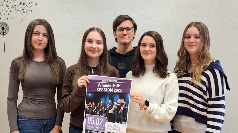 Eine Gruppe junger Menschen steht zusammen und hält ein Plakat für ein Live-Konzert, WimmerPOP Session 2026, mit dem Datum 05.02. Sie wirken aufgeregt und sind lässig gekleidet.