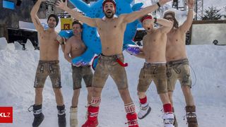 Fünf Männer posieren für ein Foto in einem verschneiten Gebiet, alle in traditioneller deutscher Kleidung. Sie haben ihre Arme erhoben und scheinen Spaß zu haben. Hinter ihnen steht ein großes blaues Aufblasbares.