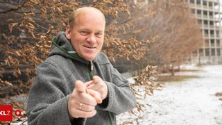 Ein Mann in einer grauen Kapuzenjacke mit grünem Reißverschluss steht in einem verschneiten Bereich und macht eine Handgeste.