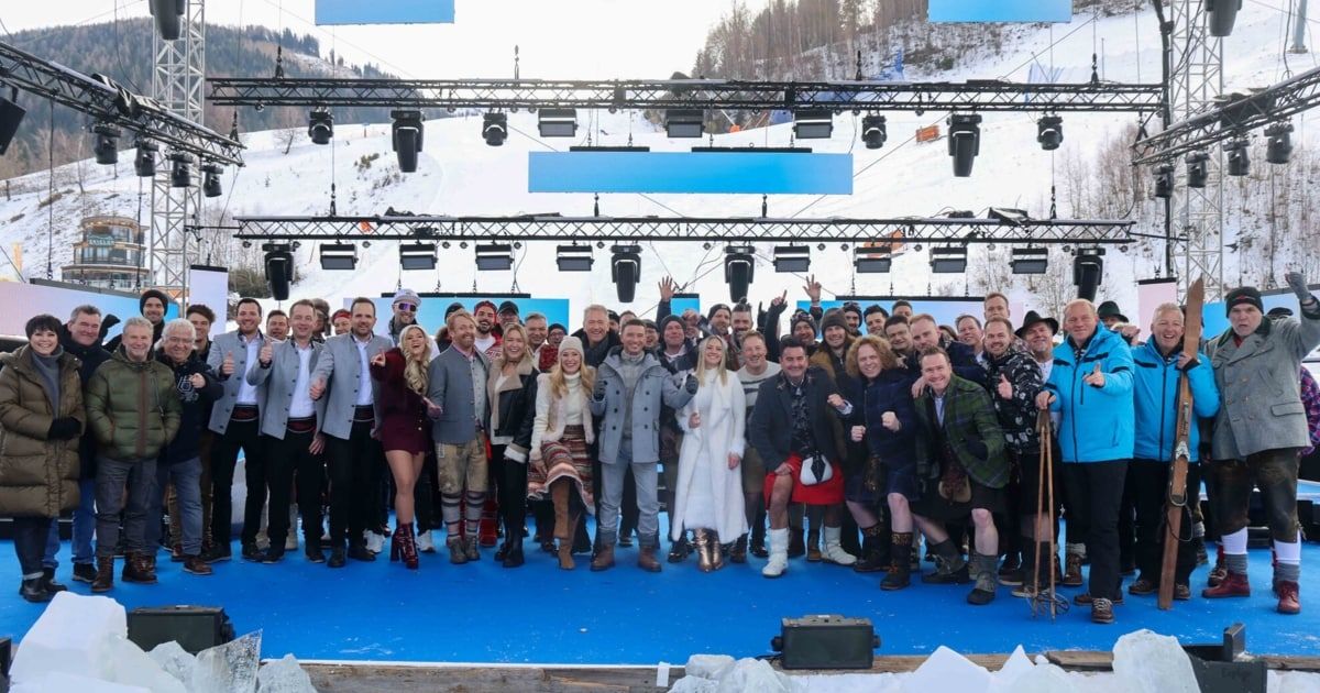 Eine Gruppe von Menschen steht auf einer Bühne in einem verschneiten Bereich, trägt Winterkleidung und posiert für ein Foto. Scheinwerfer und ein blauer Hintergrund befinden sich hinter ihnen.