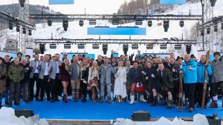 Eine Gruppe von Menschen steht auf einer Bühne in einem verschneiten Bereich, trägt Winterkleidung und posiert für ein Foto. Scheinwerfer und ein blauer Hintergrund befinden sich hinter ihnen.
