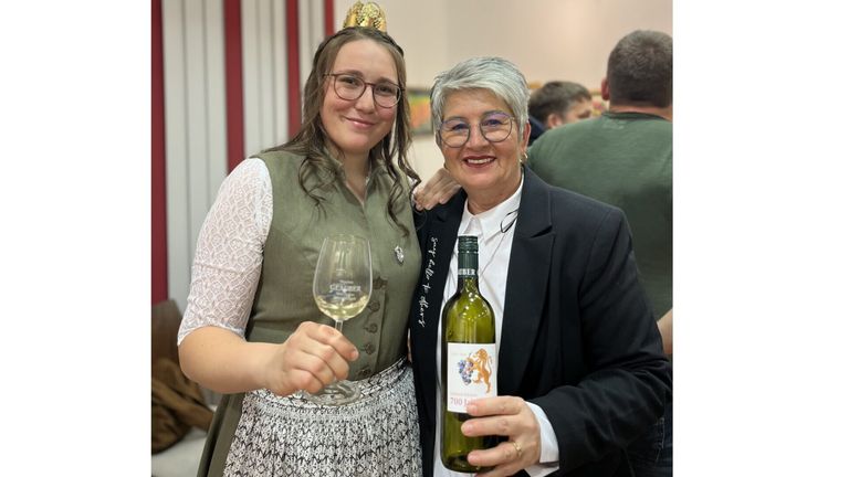 Zwei Frauen lächeln und halten eine Weinflasche und ein Weinglas. Die Frau auf der linken Seite trägt eine Krone und eine grüne Weste. Die Frau auf der rechten Seite trägt einen schwarzen Anzug und hält eine Flasche Wein.