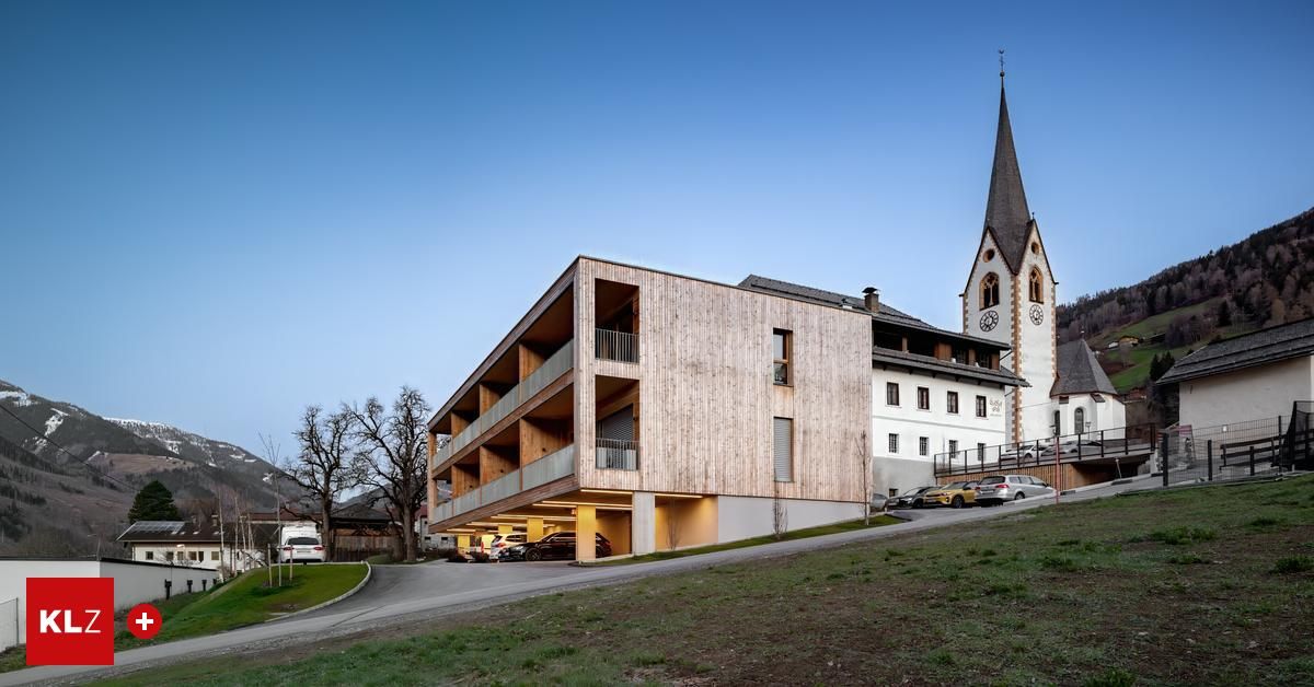 Ein modernes Holzgebäude mit mehreren Balkonen und einer Kirche mit einem Turm im Hintergrund. Mehrere Autos sind vor dem Gebäude geparkt.