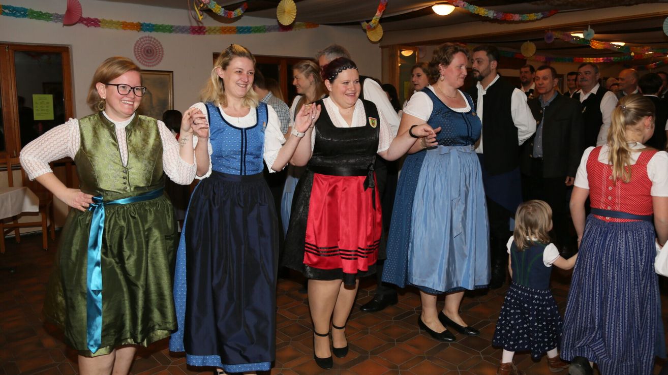 Mehrere Frauen in traditionellen Kleidern tanzen zusammen in einem Raum mit gefliesten Böden, während andere zusehen. Bunte Dekorationen hängen von der Decke herab.