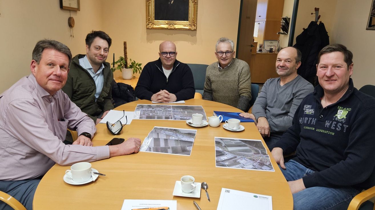 Fünf Männer sitzen um einen Tisch mit Architekturzeichnungen, Tassen und Stiften. Sie lächeln und scheinen eine Besprechung zu führen. Ein Bilderrahmen und eine Topfpflanze sind im Hintergrund.