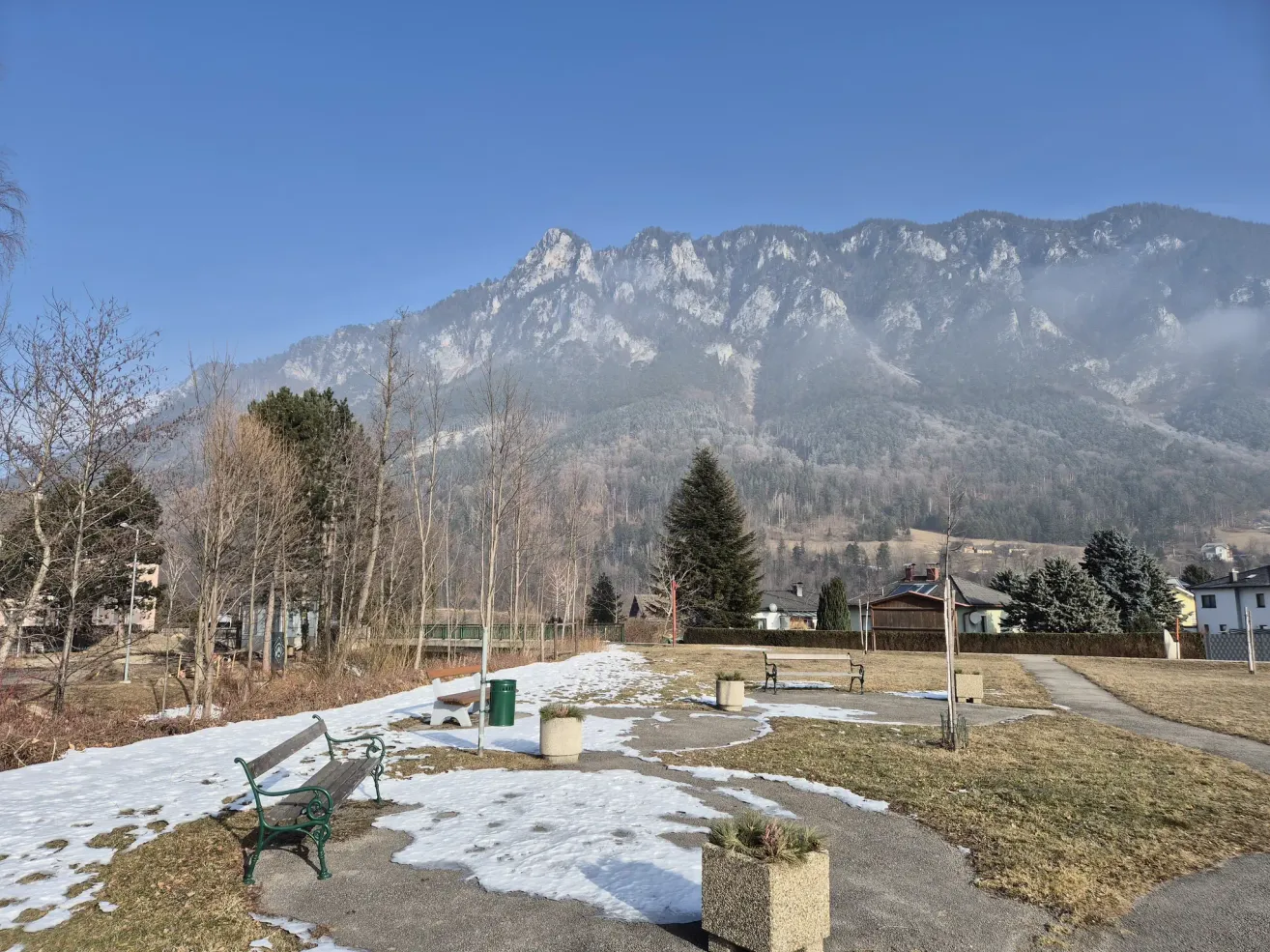 Ein Park mit Schnee auf dem Boden und Bänken, umgeben von Bäumen und Bergen im Hintergrund.