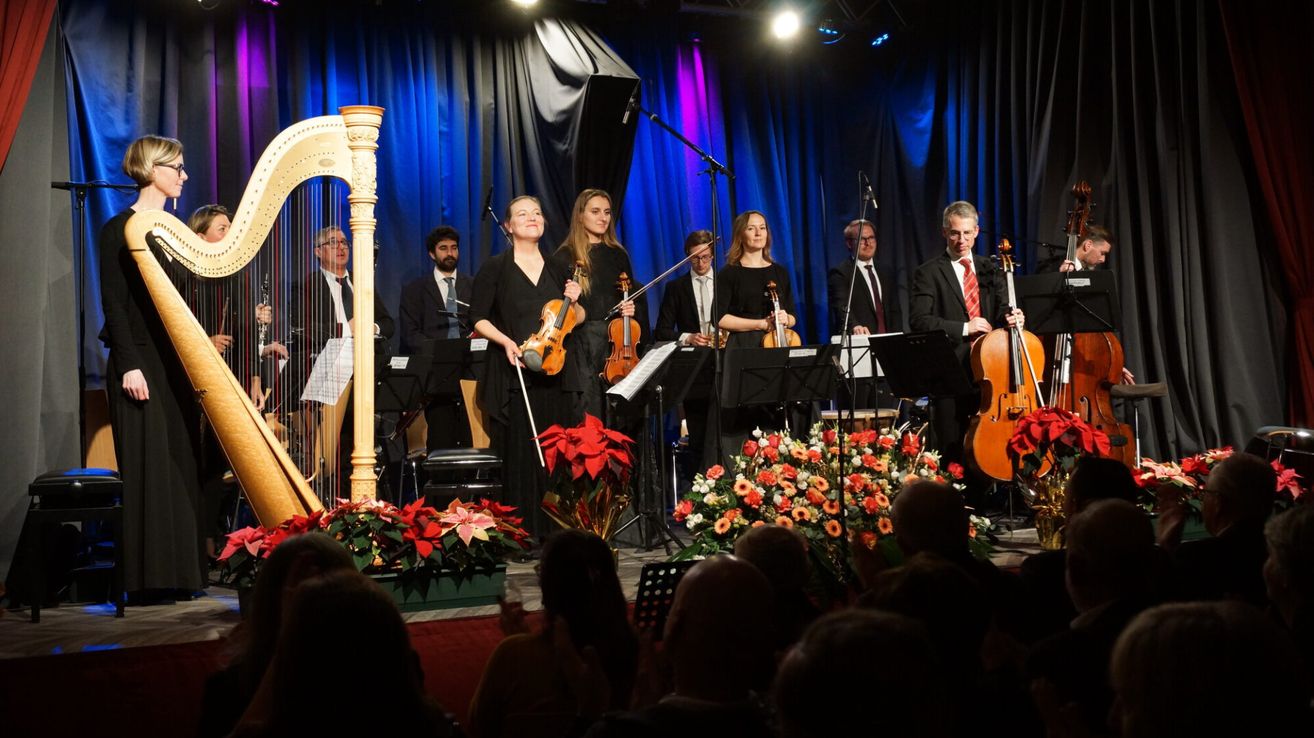 Ein Musikensemble tritt auf der Bühne auf, bestehend aus Geigern und einem Harfenisten, mit roten Blumen im Vordergrund und einem Publikum im Hintergrund.