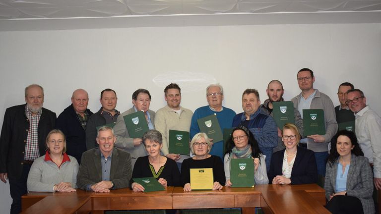 Eine Gruppe von Erwachsenen steht und sitzt hinter einem Holztisch und posiert für ein Foto mit Zertifikaten. Sie tragen formelle Kleidung und die Zertifikate sind grün. Eine weiße Wand ist hinter ihnen.