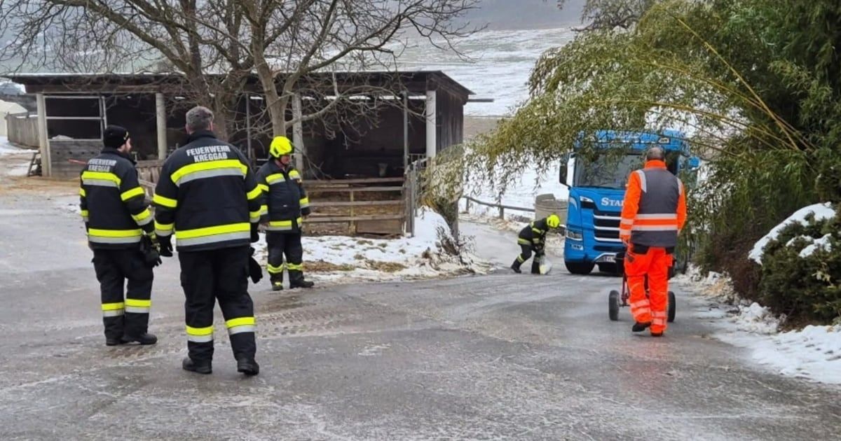 Eine Gruppe von Feuerwehrleuten steht auf einer vereisten Straße. Ein Feuerwehrmann hockt sich neben eine Barriere, und ein blauer Lastwagen ist in der Nähe. Bäume und ein Schuppen sind im Hintergrund.