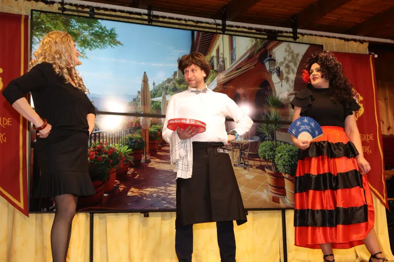 Drei Personen auf der Bühne mit einem malerischen Hintergrund einer spanischen Stadt, ein Mann hält eine rote Box, und zwei Frauen tragen schwarze und rote Kleider.