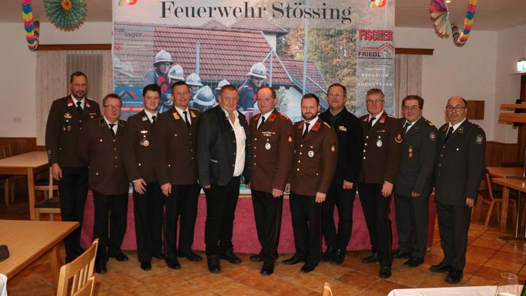Eine Gruppe von Feuerwehrleuten in Uniform posiert für ein Foto vor einem Banner. Das Banner zeigt Bilder von Feuerwehrleuten und einem Haus.