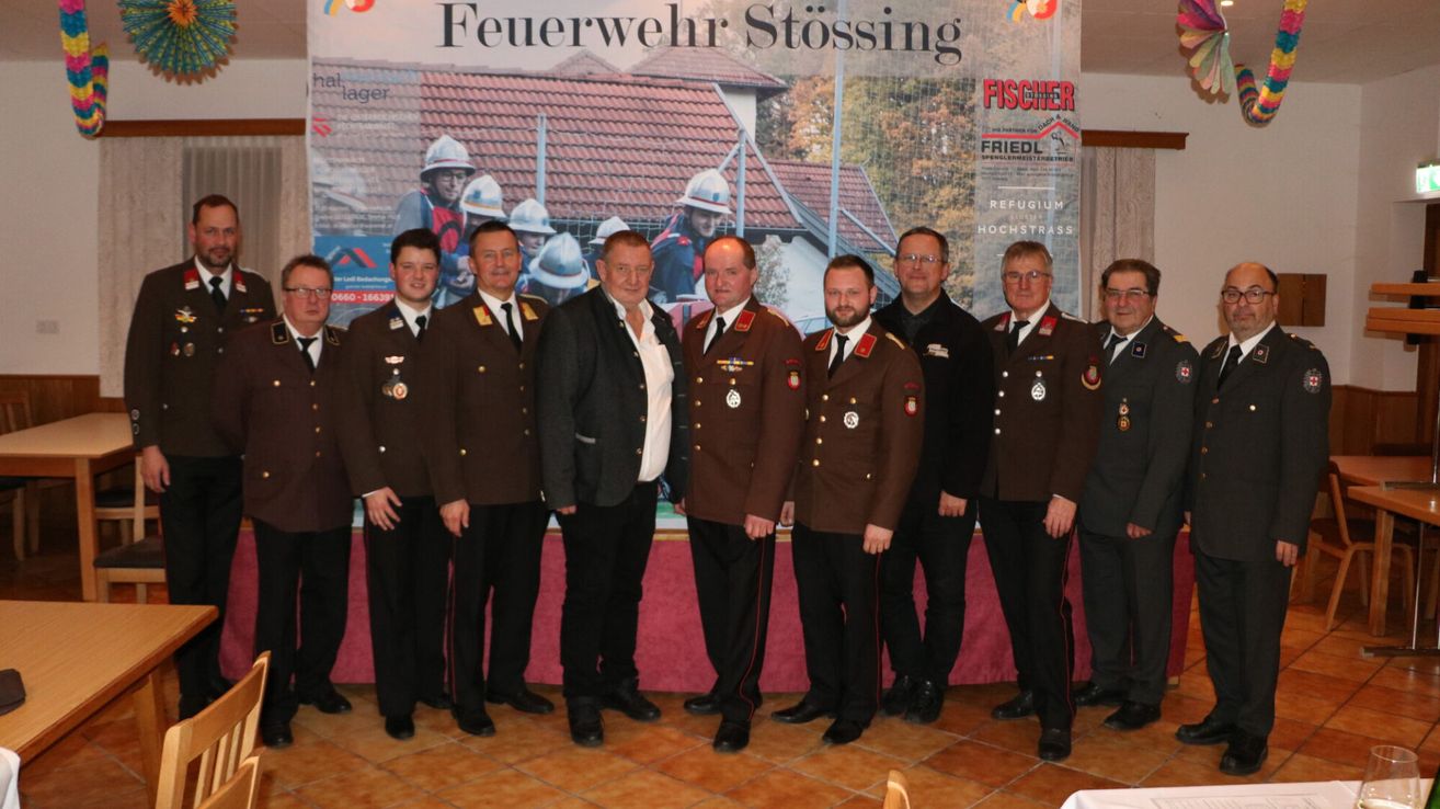 Eine Gruppe von Feuerwehrleuten in Uniform posiert für ein Foto vor einem Banner. Das Banner zeigt Bilder von Feuerwehrleuten und einem Haus.
