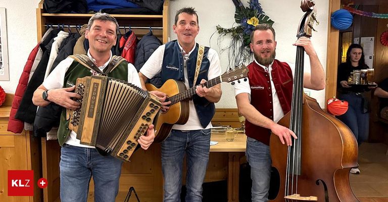 Drei Männer, lächelnd, stehen in einem Raum. Einer hält ein Akkordeon, ein anderer eine Gitarre und der dritte einen Bass. Hinter ihnen hängt eine Wand mit Blumen und Jacken an einem Ständer.