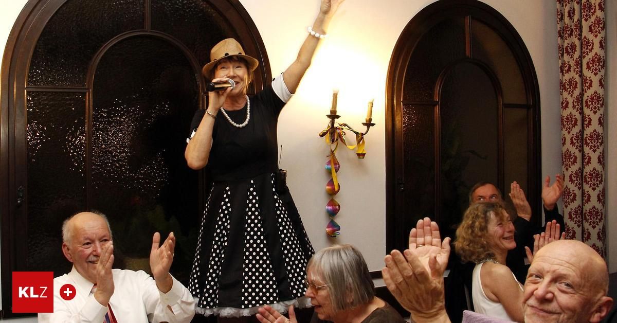 Eine Frau singt in ein Mikrofon auf einer Party. Sie trägt ein schwarzes Tupfenkleid und einen Hut. Das Publikum klatscht. Die Wand hat einen dekorativen Kerzenhalter.