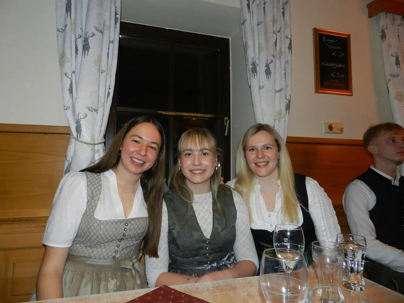 Drei Frauen sitzen lächelnd an einem Tisch mit Weingläsern und einer Speisekarte vor ihnen. Sie tragen traditionelle Kleidung und sind in einem Raum mit einem Fenster und Vorhängen. Eine Tafel mit Text ist an der Wand montiert.