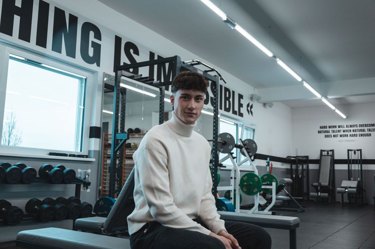 A man in a gym, wearing a white turtleneck sweater, is sitting on a bench and smiling. Behind him are various gym equipment, and the wall has a motivational quote.
