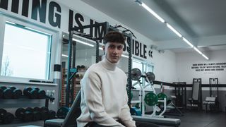 A man in a gym, wearing a white turtleneck sweater, is sitting on a bench and smiling. Behind him are various gym equipment, and the wall has a motivational quote.