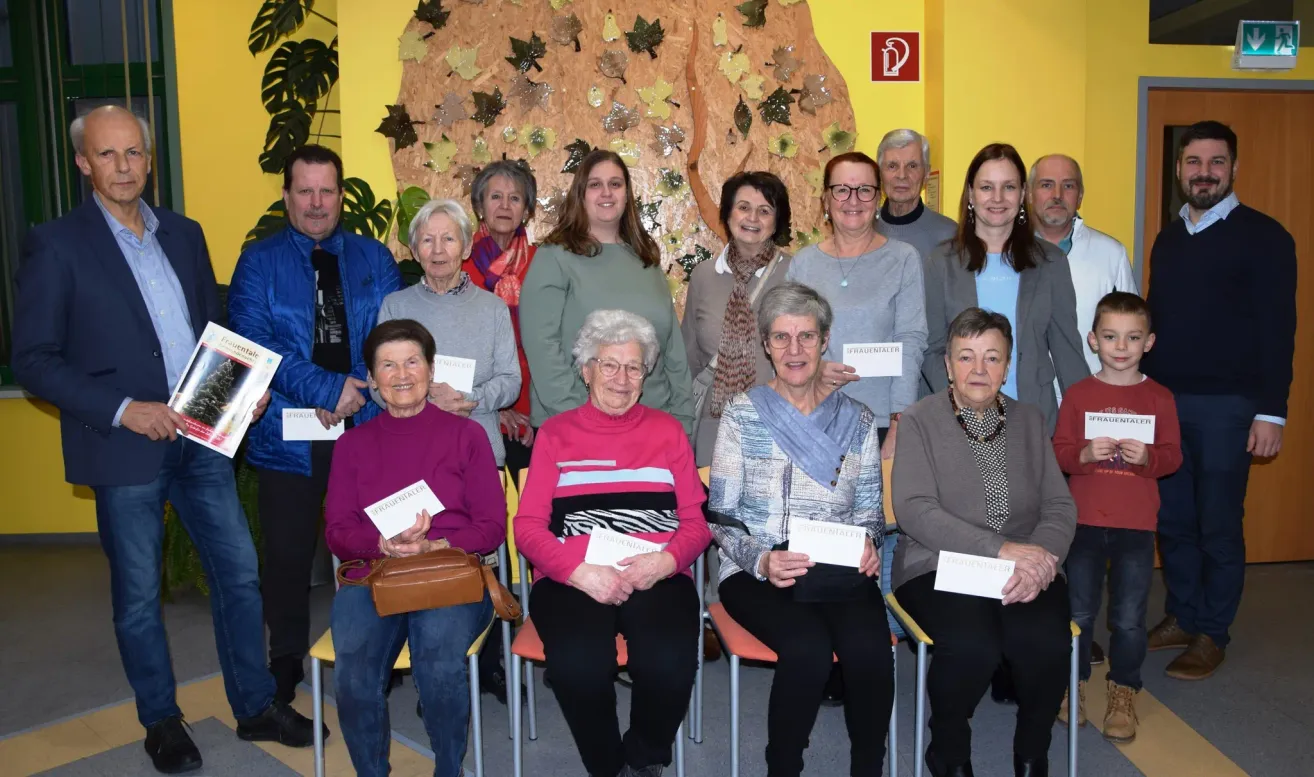 A group of elderly people pose for a photo, holding certificates. They are seated on chairs, with some standing behind them. They are all smiling.