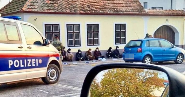 A police vehicle is parked, and a blue car is driving by a building. Several people are sitting on the ground, and a man is standing.