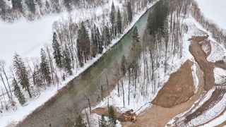 An aerial view of a snowy landscape with a river, dense trees, and a yellow machinery vehicle digging soil on the right.