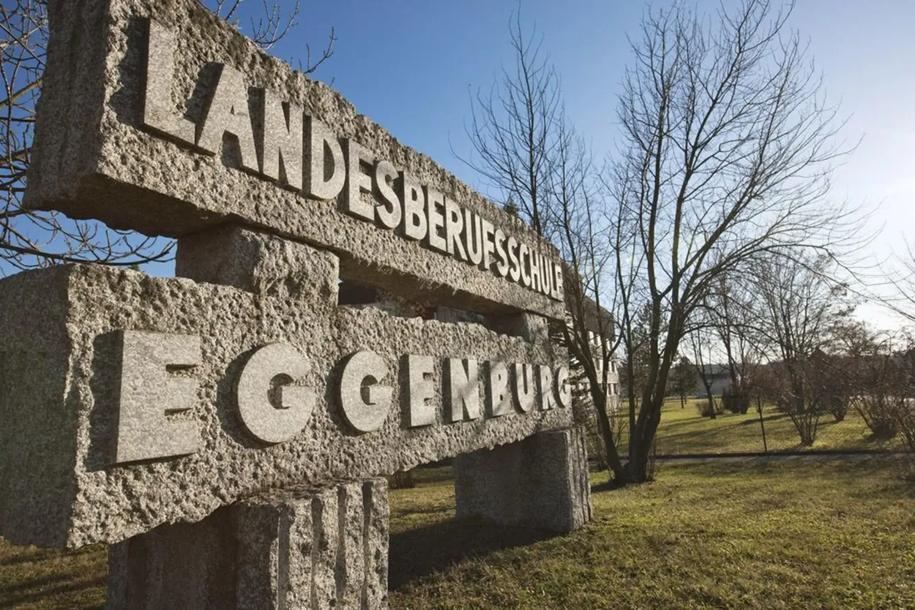 A stone sign with the words 'Landesberufsschule' and 'Egensburg' in front of a grassy field with a tree and clear sky.