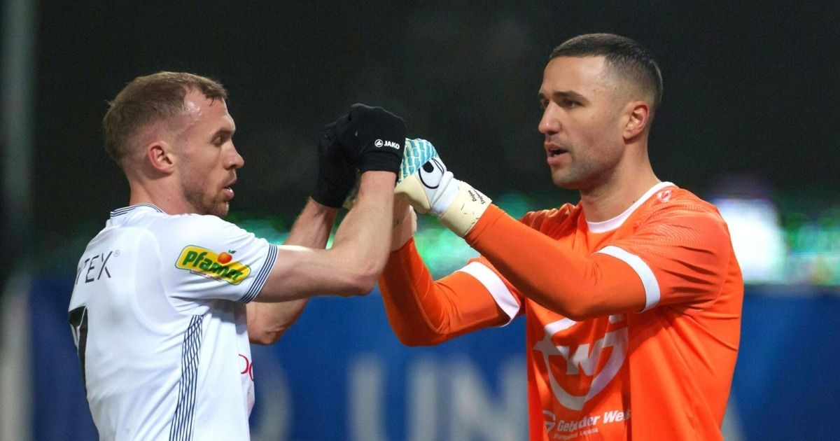 Two soccer players, one in white and one in orange, shake hands in a stadium.