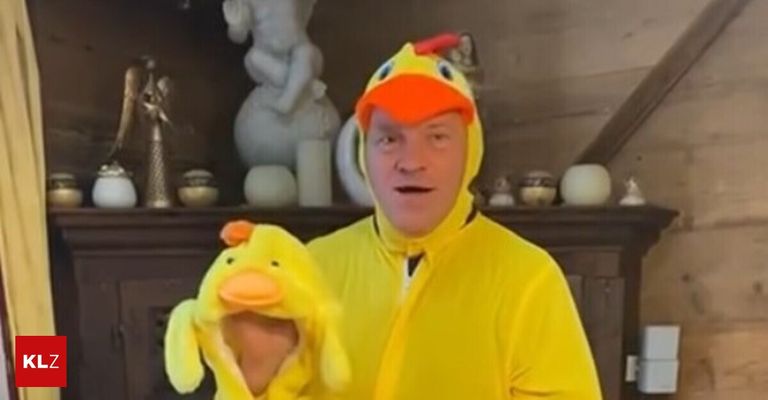 A man dressed in a yellow duck costume with an orange beak and a hat is standing in front of a shelf with decorative items.