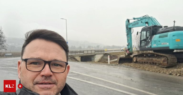 A man with glasses stands near a road with a construction vehicle in the background.