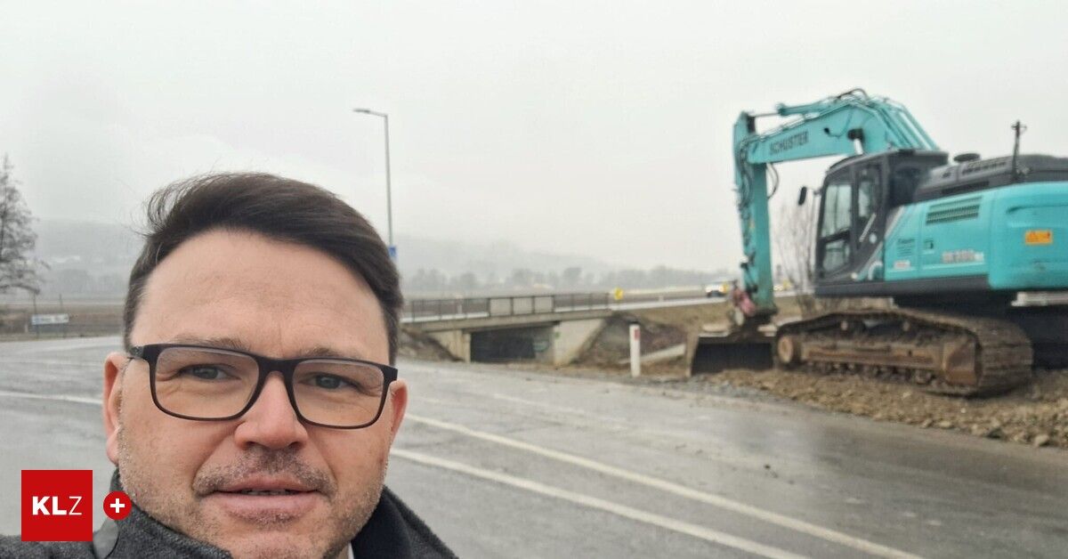 A man with glasses stands near a road with a construction vehicle in the background.