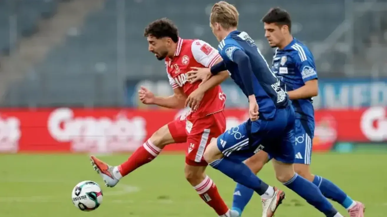 Two soccer players in red and two in blue are playing on the field. One player in red is kicking the ball while another player in blue is trying to block him.