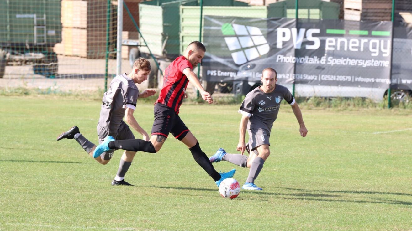 Three men are playing soccer on a grassy field. One man in a red and black jersey is kicking the ball. The other two men are in gray jerseys, one defending and the other running.