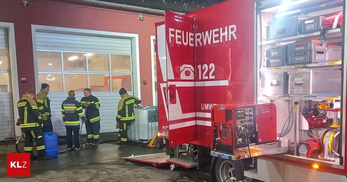 Three firefighters in uniform stand outside a fire station, one holding a hose near a fire truck labeled 'Feuerwehr 122'. The truck has a red body with white markings and a generator on the back.