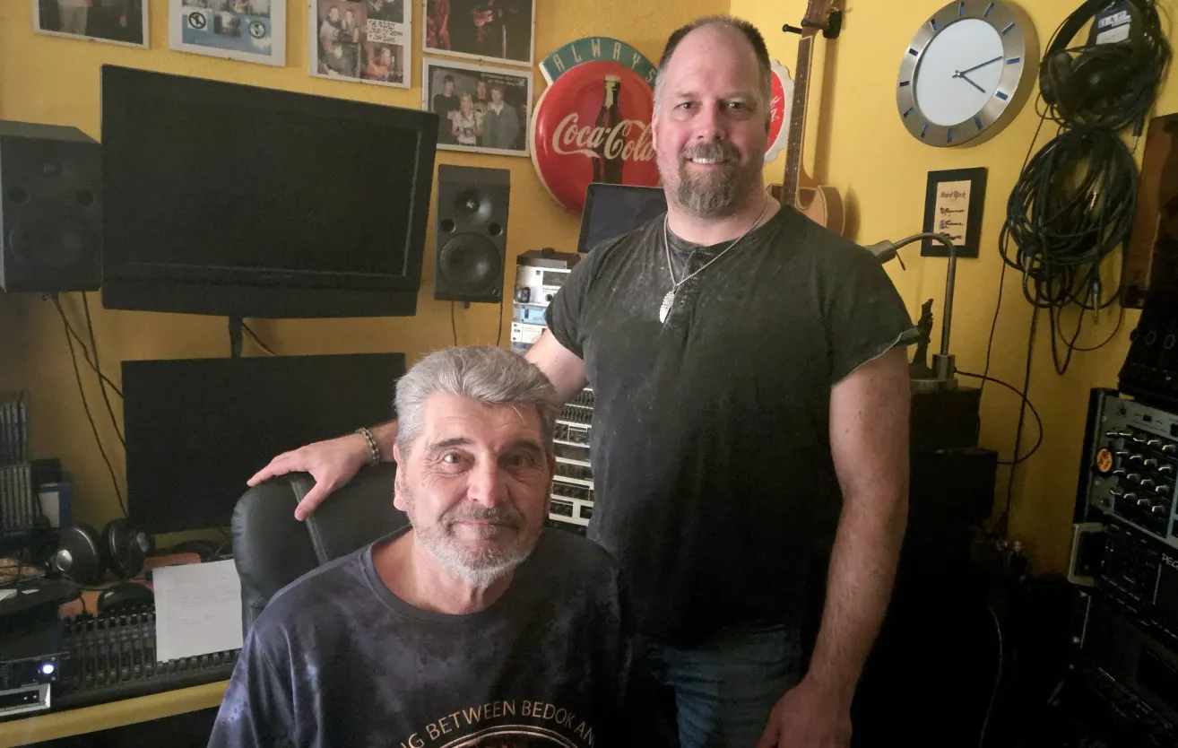 Two men are standing in a room with yellow walls. One is sitting in a chair, and the other is standing behind him. There are guitars and a Coca-Cola sign on the wall.