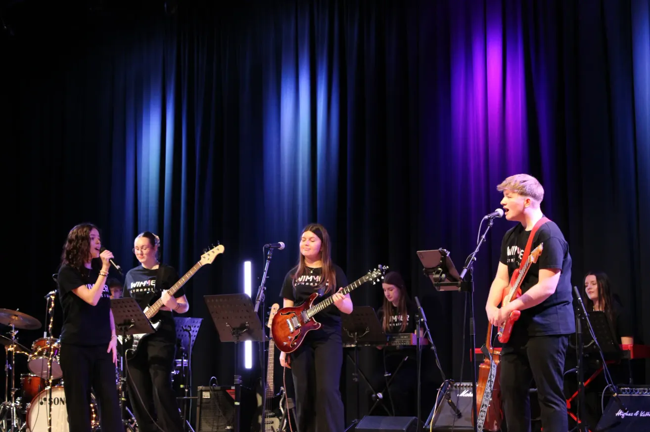 Eine Band tritt auf der Bühne auf, mit lila und blauen Lichtern im Hintergrund. Der Leadsänger singt, während die Gitarristen hinter ihm spielen.