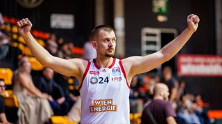 A basketball player celebrates with his arms raised at a sports event. He wears a white jersey with 'WIEN ENERGIE' and the number '24'. The audience watches from the stands.