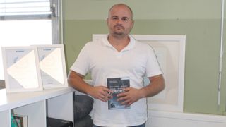 A man in a white polo shirt is holding a book with both hands in front of his body. The book's title is Schmerz-Angst-Panik-Burnout-Verstehen.