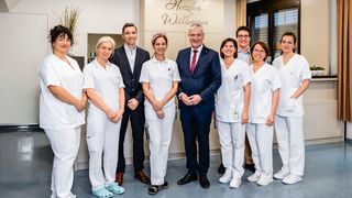 A group of people dressed in white lab coats and shoes, standing next to a man in a suit, in a room with a sign reading Herzlich Willkommen.
