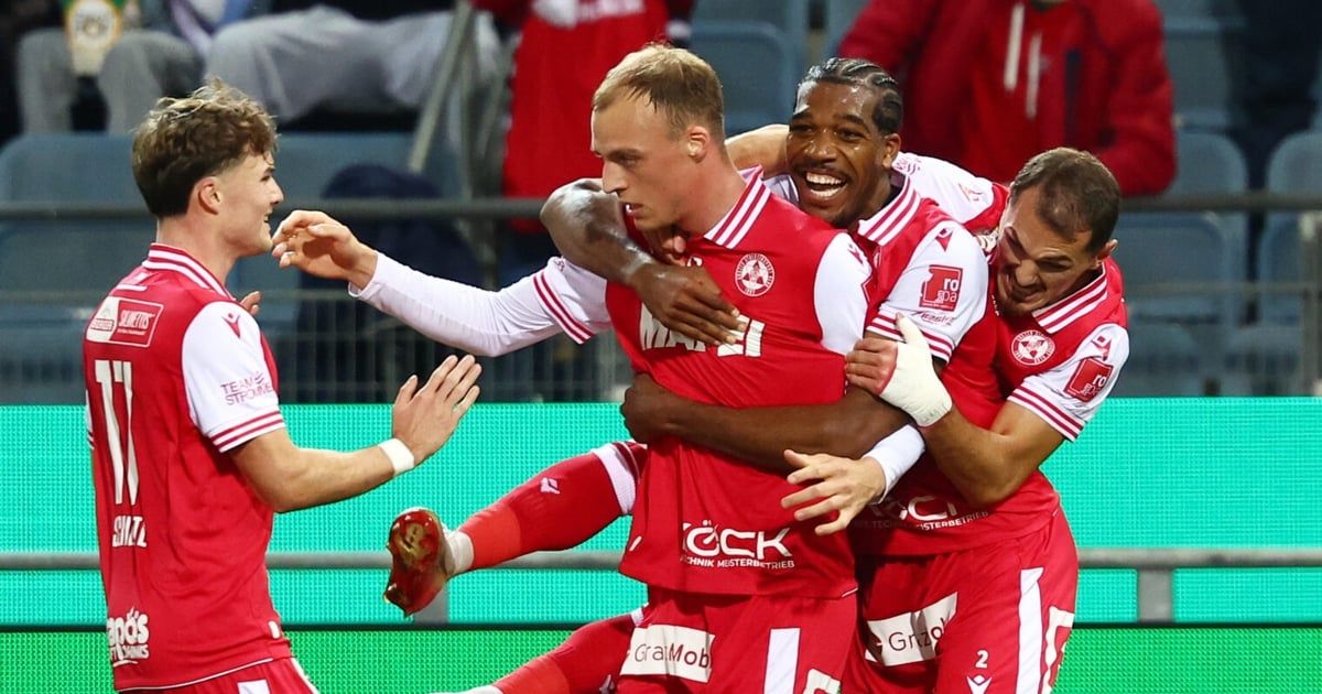 Soccer players in red uniforms celebrate a goal, surrounded by teammates and spectators, with one player showing a bandaged arm.