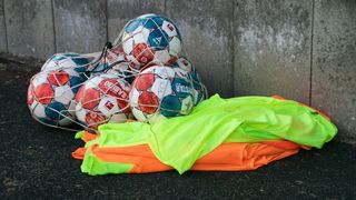 A collection of soccer balls in various designs, including Derby Star and Select, is placed in a net with reflective shirts underneath a concrete wall.