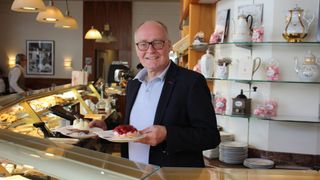 A man in a suit is standing in a bakery, smiling and holding two plates with desserts. Behind him, there are shelves with decorative items and a table with various equipment.