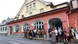 A group of people stand in front of the Kunsthaus building, engaging in conversation. The building has arched windows and is painted in pink and yellow.