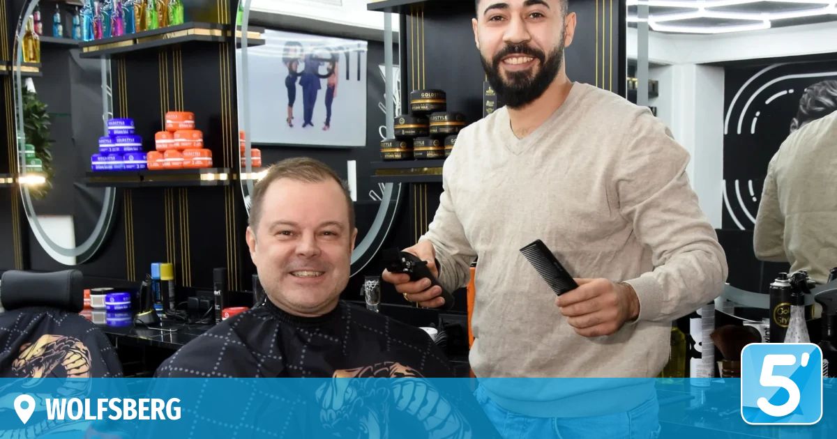 A man in a barber shop, getting his hair cut by a smiling barber holding a comb and a remote control.