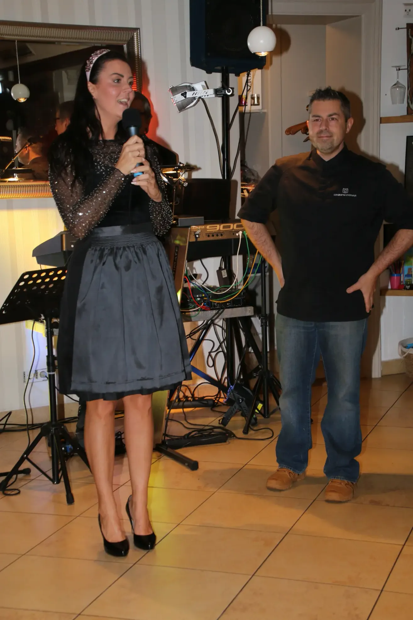 A woman in a black dress holds a microphone, possibly at an event. A man in a black shirt stands next to her, looking at her. Behind them is a table with electronic equipment.