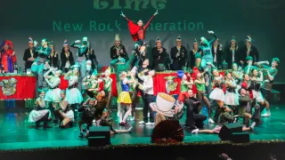 A vibrant stage performance with a group of people dressed in colorful costumes and masks. Some are dancing, others are performing acrobatics, and a few are kneeling on the stage. Behind them, a red curtain is draped on the stage.
