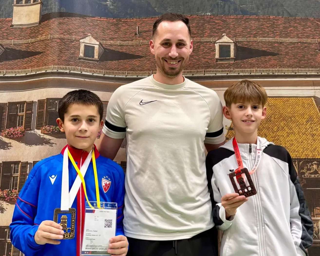 A smiling man in a white shirt stands between two young boys, both wearing medals. The boy on the left has a blue jacket with a red and yellow ribbon. The boy on the right has a white jacket with a red ribbon. They are standing in front of a backdrop of a house with a brick roof.
