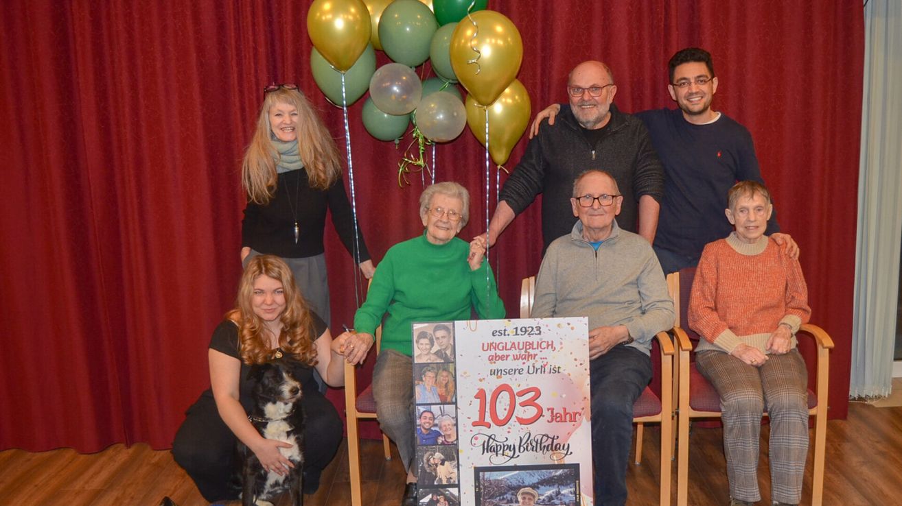 A group of people is gathered around a woman celebrating her 103rd birthday. Balloons are behind them. The woman is holding a sign that says 103 years and Happy Birthday.