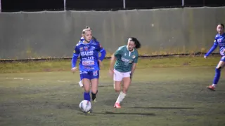 Two women play soccer on a field. One in blue, the other in green. The blue-clad woman is kicking the ball while the green-clad woman runs towards her.